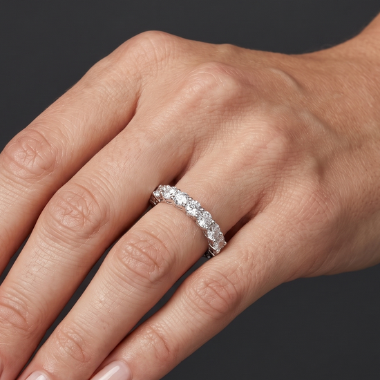 Close-up photo of a woman's hand wearing a luxurious round-cut diamond eternity band. Soft lighting highlights the sparkling ring and natural skin texture against a dark gray background.