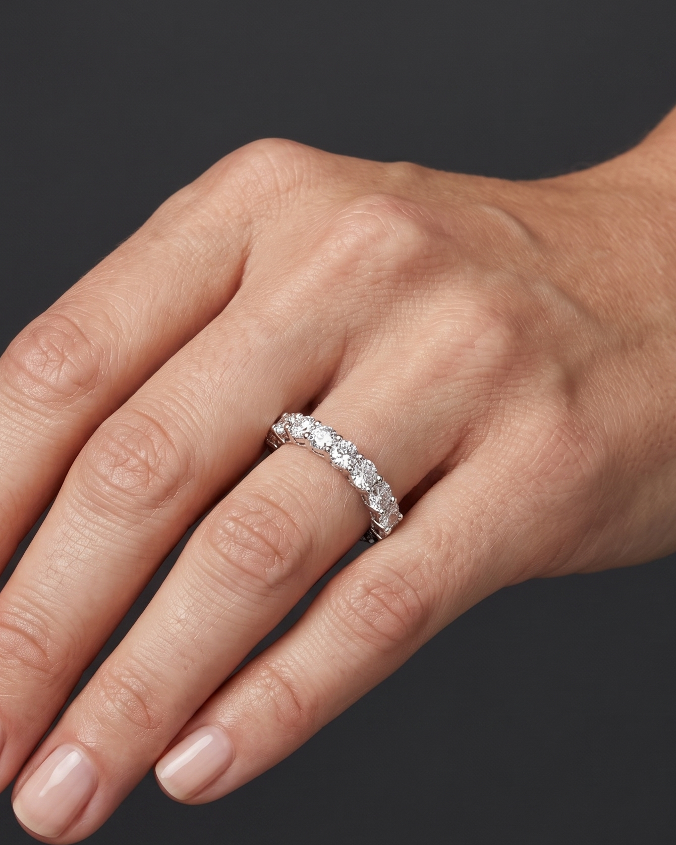 Close-up photo of a woman's hand wearing a luxurious round-cut diamond eternity band. Soft lighting highlights the sparkling ring and natural skin texture against a dark gray background.