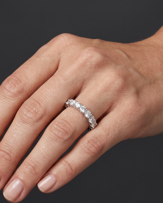 Close-up photo of a woman's hand wearing a luxurious round-cut diamond eternity band. Soft lighting highlights the sparkling ring and natural skin texture against a dark gray background.