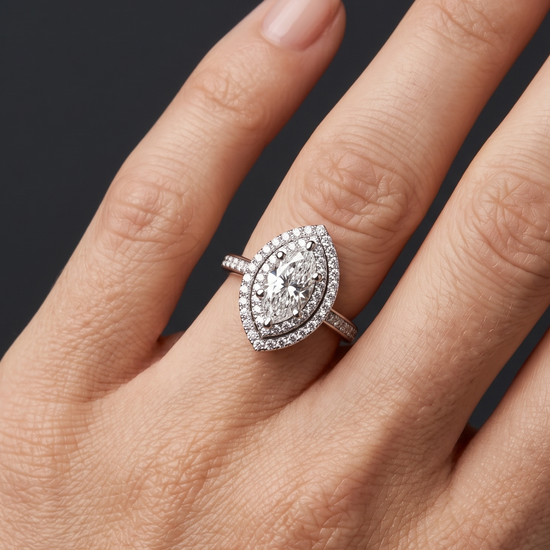 A close-up of a woman's hand elegantly displaying a large marquise-cut diamond ring with a double halo and a diamond-encrusted band. The ring rests naturally on her ring finger against a smooth, dark charcoal gray background. Soft, diffused lighting highlights the sparkling details of the jewelry and the natural texture of her skin, creating a luxurious and premium feel.