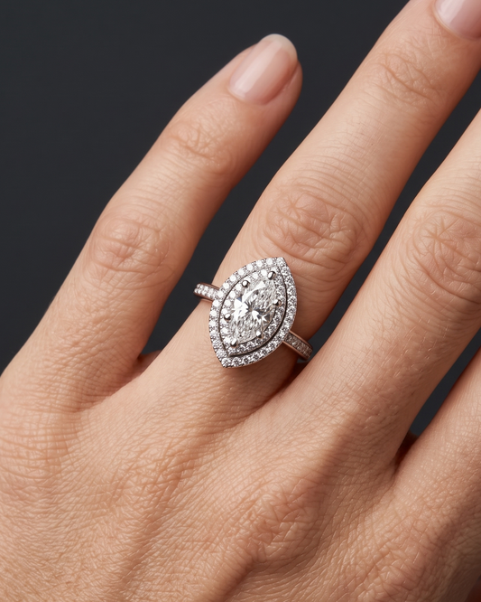 A close-up of a woman's hand elegantly displaying a large marquise-cut diamond ring with a double halo and a diamond-encrusted band. The ring rests naturally on her ring finger against a smooth, dark charcoal gray background. Soft, diffused lighting highlights the sparkling details of the jewelry and the natural texture of her skin, creating a luxurious and premium feel.