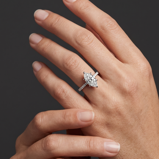 A close-up editorial photograph focusing on a woman's elegantly posed hands resting against a smooth, dark charcoal gray background. On her left ring finger, she wears a brilliant white gold or platinum engagement ring featuring a prominent marquise-cut center diamond secured by six prongs, set on a slender band lined with smaller pavé diamonds.