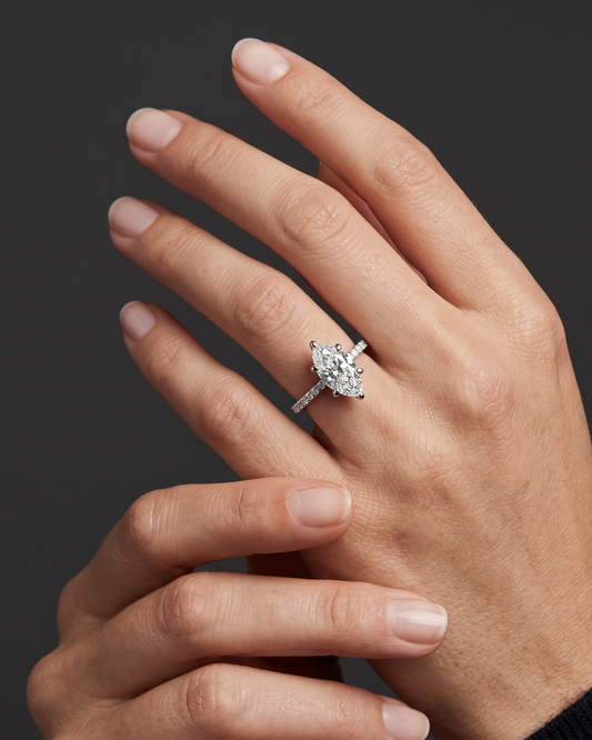A close-up editorial photograph focusing on a woman's elegantly posed hands resting against a smooth, dark charcoal gray background. On her left ring finger, she wears a brilliant white gold or platinum engagement ring featuring a prominent marquise-cut center diamond secured by six prongs, set on a slender band lined with smaller pavé diamonds.
