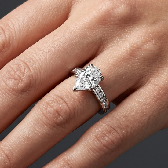 A close-up of a woman's hand resting elegantly against a dark charcoal gray background, wearing a platinum engagement ring with a large pear-cut diamond and a channel-set diamond band.