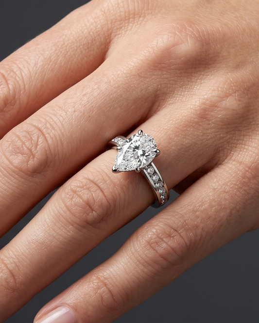 A close-up of a woman's hand resting elegantly against a dark charcoal gray background, wearing a platinum engagement ring with a large pear-cut diamond and a channel-set diamond band.