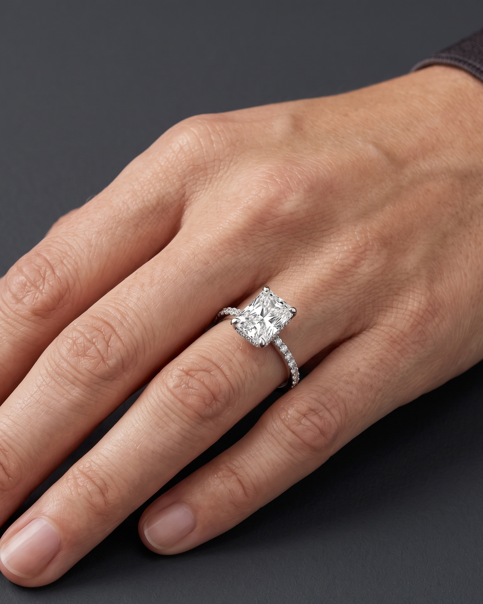 Close-up editorial shot of a woman's hand resting elegantly against a smooth, dark charcoal gray background. She is wearing a luxurious silver-toned engagement ring on her ring finger, featuring a large radiant-cut center diamond and a delicate band encrusted with smaller pavé diamonds. A slight hint of a dark long sleeve is visible at her wrist, and soft studio lighting highlights the sparkle of the jewelry and the natural texture of her skin.