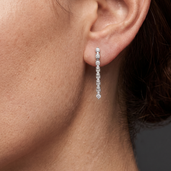 A close-up profile shot of a woman's ear and jawline showcasing a petite, linear diamond drop earring. The delicate earring features a straight line of alternating tiny round and square-cut diamonds set in silver-toned metal.