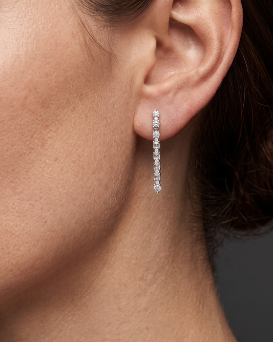 A close-up profile shot of a woman's ear and jawline showcasing a petite, linear diamond drop earring. The delicate earring features a straight line of alternating tiny round and square-cut diamonds set in silver-toned metal.