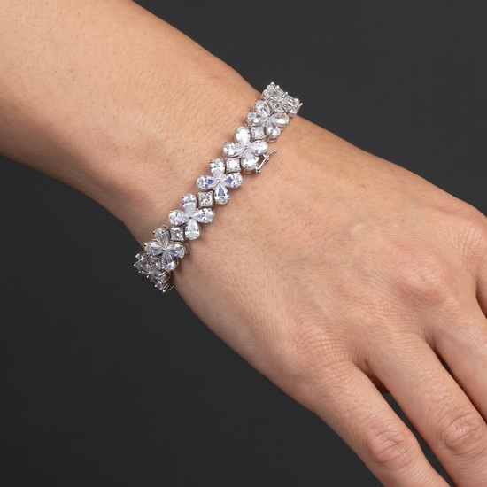 A close-up photograph of a beautifully posed hand and wrist resting against a smooth, dark charcoal gray background. The wrist is adorned with a sparkling, silver-tone bracelet made of linked floral clusters set with clear, diamond-like stones. Soft, natural lighting highlights the brilliance of the jewelry and the realistic texture of the skin.