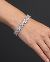 A close-up photograph of a beautifully posed hand and wrist resting against a smooth, dark charcoal gray background. The wrist is adorned with a sparkling, silver-tone bracelet made of linked floral clusters set with clear, diamond-like stones. Soft, natural lighting highlights the brilliance of the jewelry and the realistic texture of the skin.