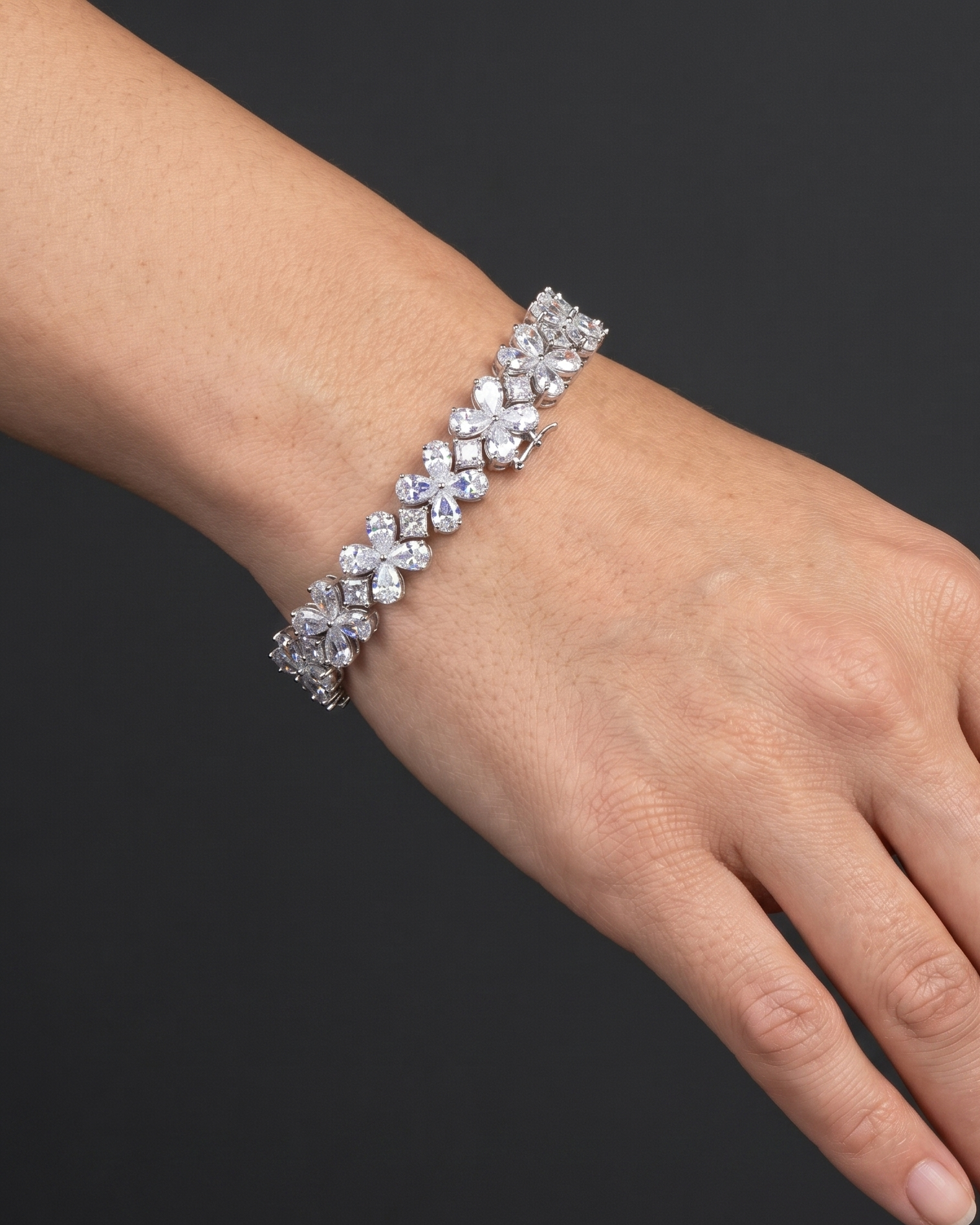 A close-up photograph of a beautifully posed hand and wrist resting against a smooth, dark charcoal gray background. The wrist is adorned with a sparkling, silver-tone bracelet made of linked floral clusters set with clear, diamond-like stones. Soft, natural lighting highlights the brilliance of the jewelry and the realistic texture of the skin.
