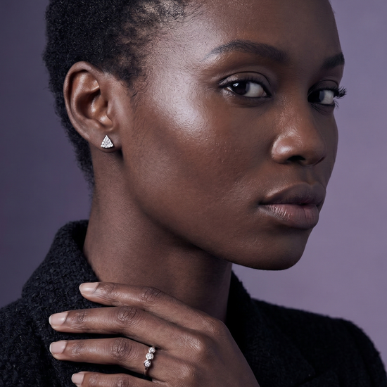 Model wearing small triangular pave diamond stud earrings and a dainty three-stone bezel ring, styled with a black tweed blazer.