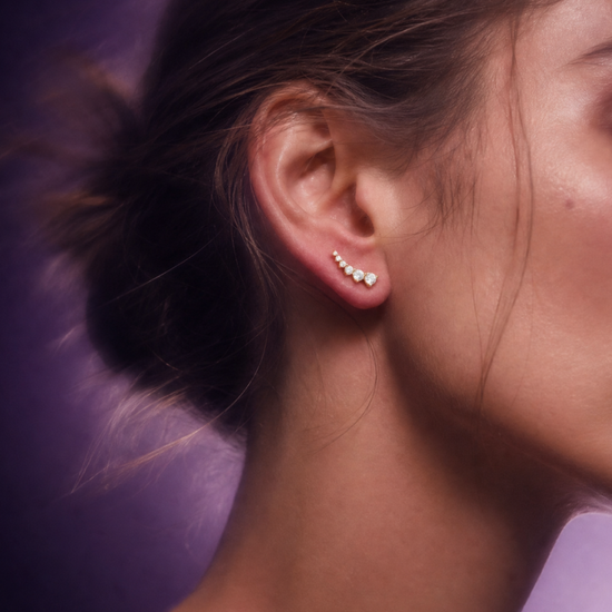 Detailed close-up view of a model's ear lobe wearing a single yellow gold and diamond ear climber earring, featuring a curved sequence of multiple brilliant-cut diamonds with a distinct top cluster, set against a moody blurred purple and lavender gradient background.