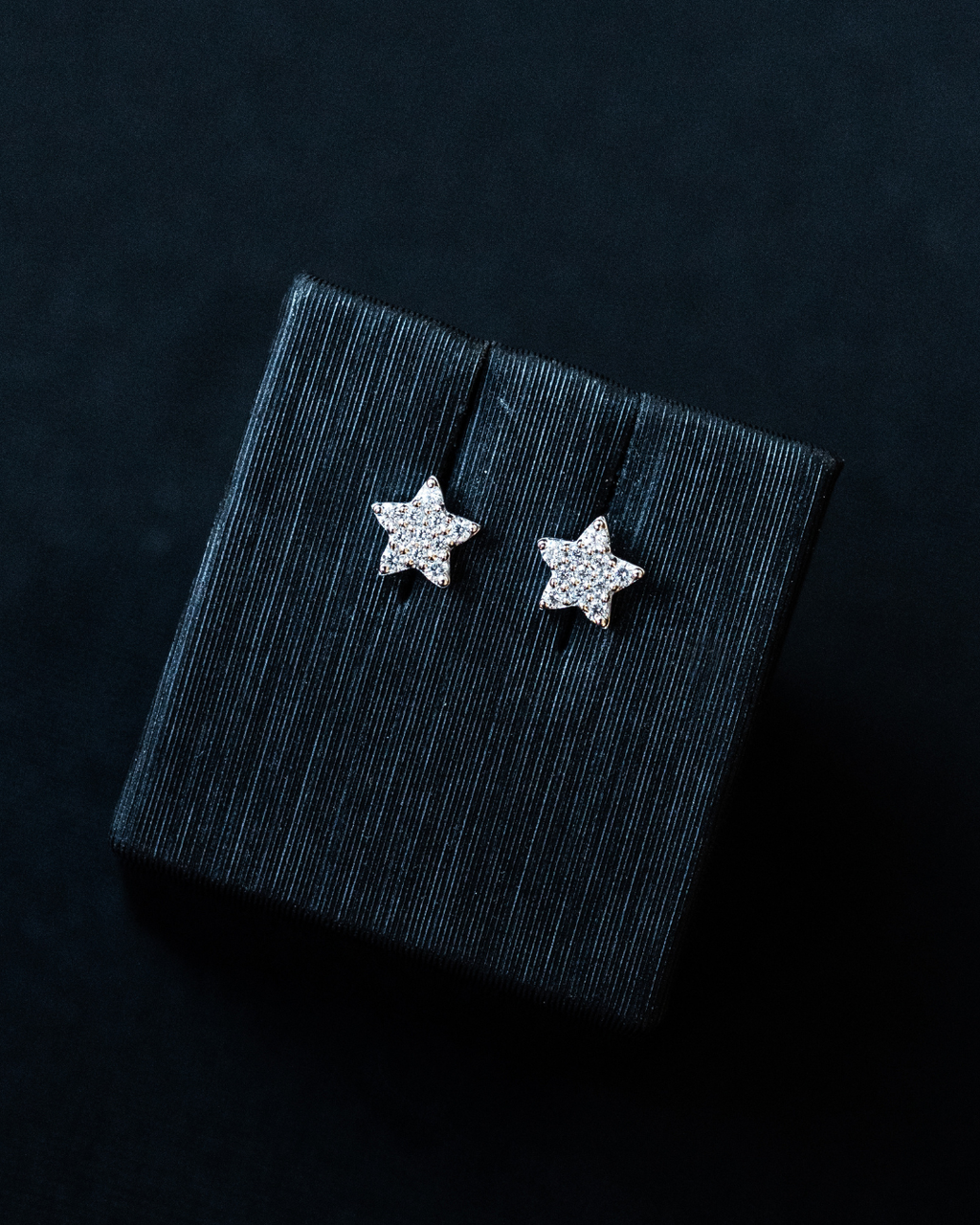 A pair of sparkling, star-shaped stud earrings displayed on a dark, textured jewelry box insert against a black background.