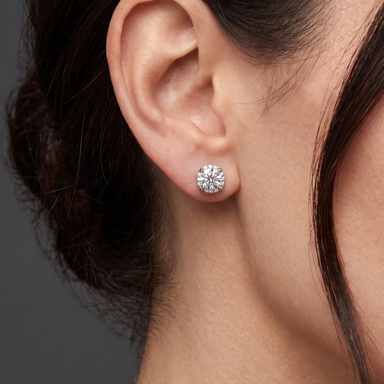 A close-up side profile shot focusing on a person's ear and jawline against a dark gray studio background. The person has dark hair pulled back and is modeling a large, sparkling round-cut diamond stud earring resting on the earlobe.