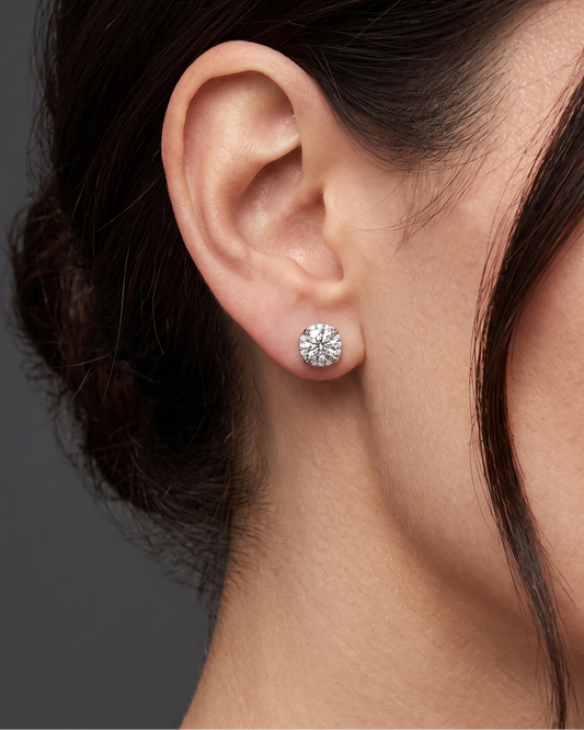 A close-up side profile shot focusing on a person's ear and jawline against a dark gray studio background. The person has dark hair pulled back and is modeling a large, sparkling round-cut diamond stud earring resting on the earlobe.