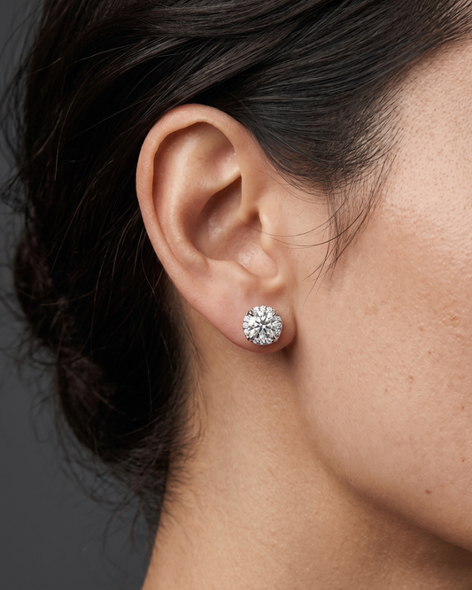 A close-up side profile shot focusing on a person's ear and jawline against a dark gray studio background. The person has dark hair pulled back and is modeling a large, sparkling round-cut diamond stud earring resting on the earlobe.