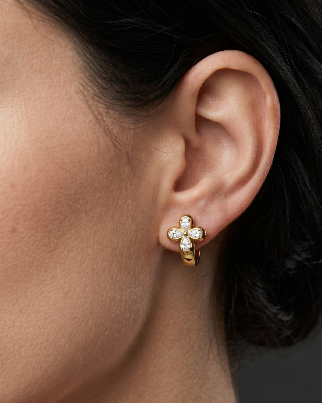 Close-up lifestyle view of a 14k yellow gold huggie earring featuring a prominent clover-shaped four-leaf flower motif studded with pear-cut white diamonds.