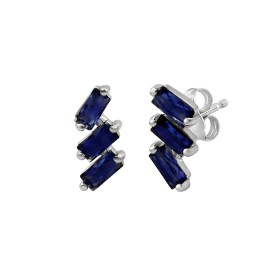 A pair of cascading stud earrings featuring a vertical arrangement of three baguette-cut blue sapphires. The rectangular gemstones are staggered in a step-like formation and held in a minimalist 18k white gold setting, shown against a clean, white studio background.