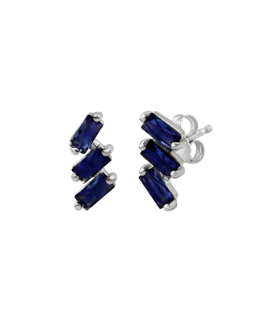A pair of cascading stud earrings featuring a vertical arrangement of three baguette-cut blue sapphires. The rectangular gemstones are staggered in a step-like formation and held in a minimalist 18k white gold setting, shown against a clean, white studio background.