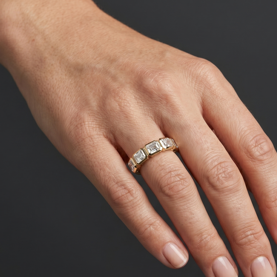 Close-up editorial photograph of a woman's hand elegantly posed against a smooth, dark charcoal gray background. She is wearing a petite gold ring on her ring finger, which features a row of five bezel-set, emerald-cut clear gemstones. Soft lighting highlights the details of the jewelry and the natural texture of her skin.