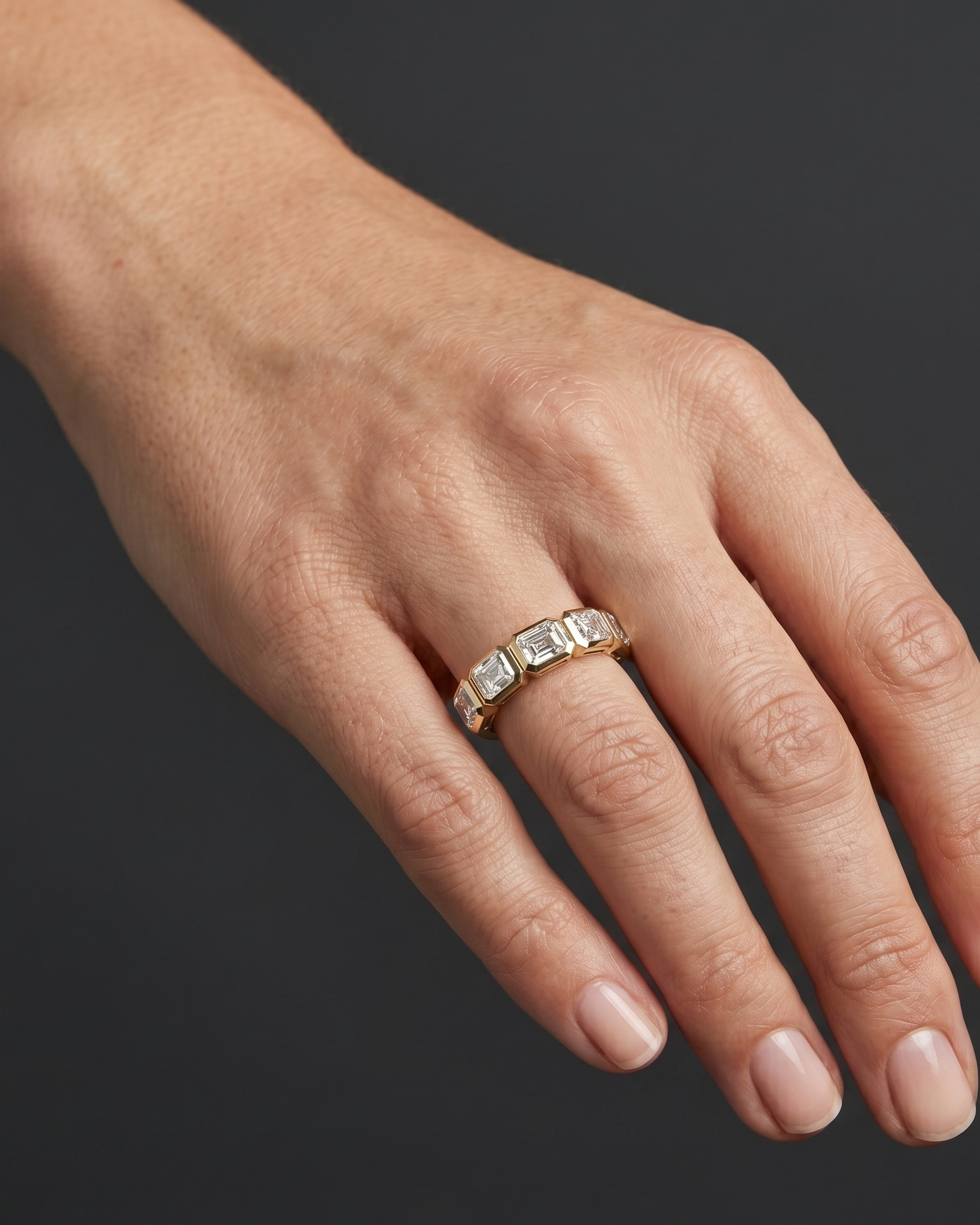 Close-up editorial photograph of a woman's hand elegantly posed against a smooth, dark charcoal gray background. She is wearing a petite gold ring on her ring finger, which features a row of five bezel-set, emerald-cut clear gemstones. Soft lighting highlights the details of the jewelry and the natural texture of her skin.