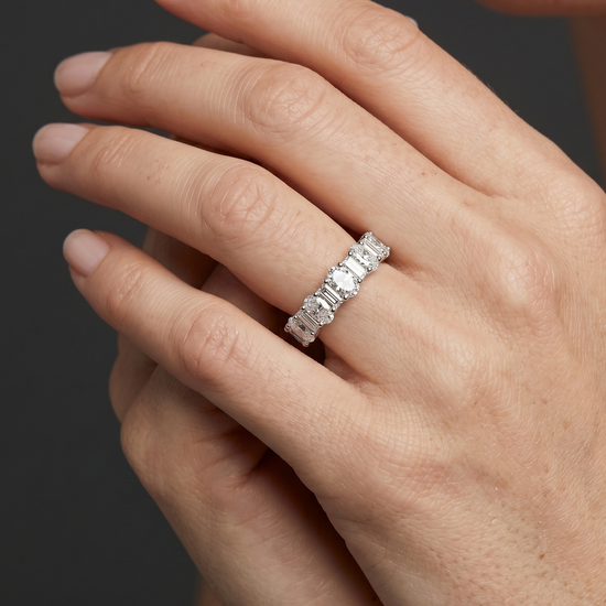 Close-up of a gracefully posed woman's hand wearing a petite, silver-toned ring featuring an alternating pattern of oval and baguette-cut diamonds. The hand, with natural skin texture and nude manicured nails, is set against a smooth, dark charcoal gray background under soft, diffused studio lighting.