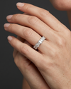 Close-up of a gracefully posed woman's hand wearing a petite, silver-toned ring featuring an alternating pattern of oval and baguette-cut diamonds. The hand, with natural skin texture and nude manicured nails, is set against a smooth, dark charcoal gray background under soft, diffused studio lighting.