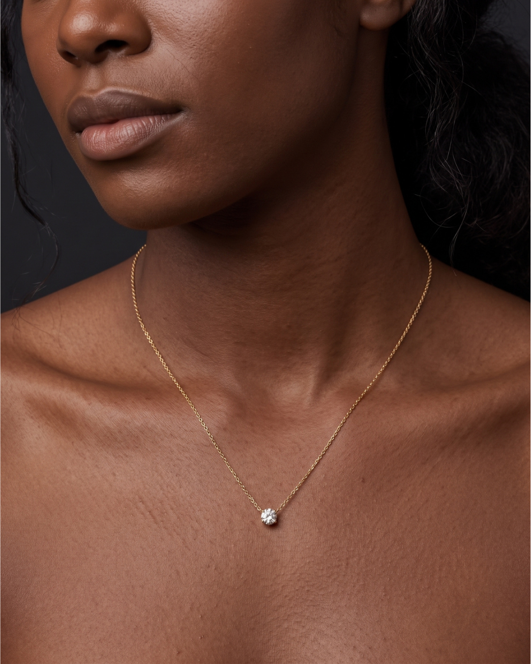 A close-up of a person's lower face and neck wearing a delicate yellow gold chain featuring a noticeably larger, single round 2-carat diamond pendant resting just below the collarbone.