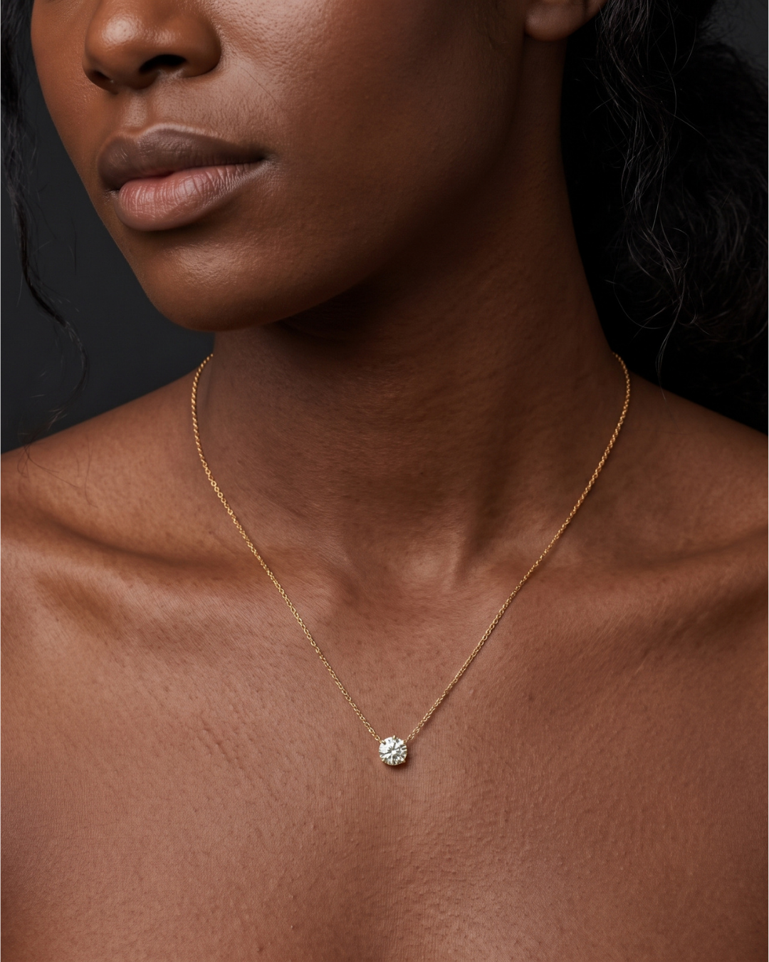 A close-up of a person's lower face and neck wearing a delicate yellow gold chain featuring a small, single round 1-carat diamond pendant resting just below the collarbone.