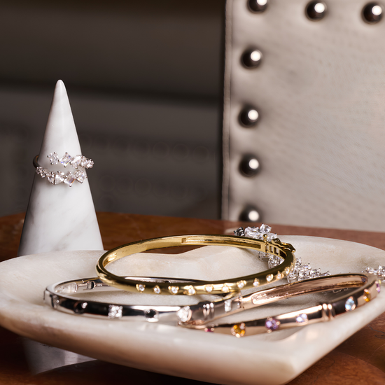 High-polish gold and silver diamond bangles arranged on a marble dish beside a diamond ring on a cone display, styled on a wooden surface with an upholstered chair in the background.