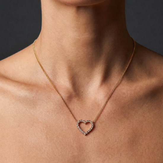 A close-up of a model's neck and upper chest wearing a delicate gold chain necklace with an open heart-shaped pendant set with small clear stones, against a dark gray background.