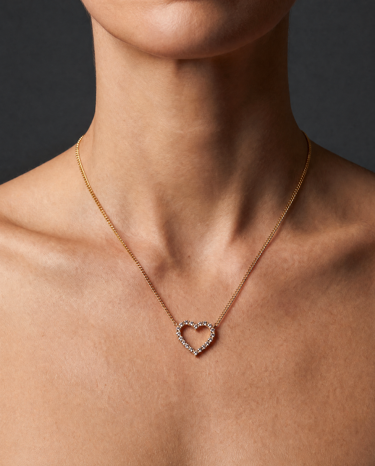 A close-up of a model's neck and upper chest wearing a delicate gold chain necklace with an open heart-shaped pendant set with small clear stones, against a dark gray background.