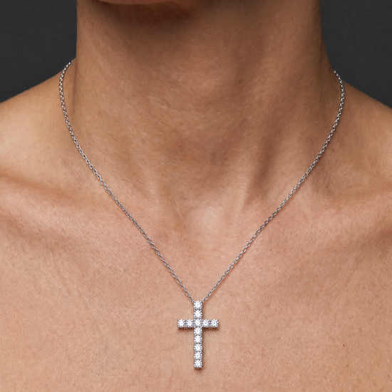 A silver cross pendant necklace adorned with small clear stones, worn on a model's neck against a dark gray background.