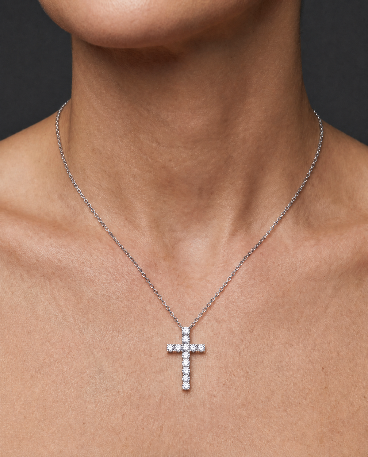 A silver cross pendant necklace adorned with small clear stones, worn on a model's neck against a dark gray background.