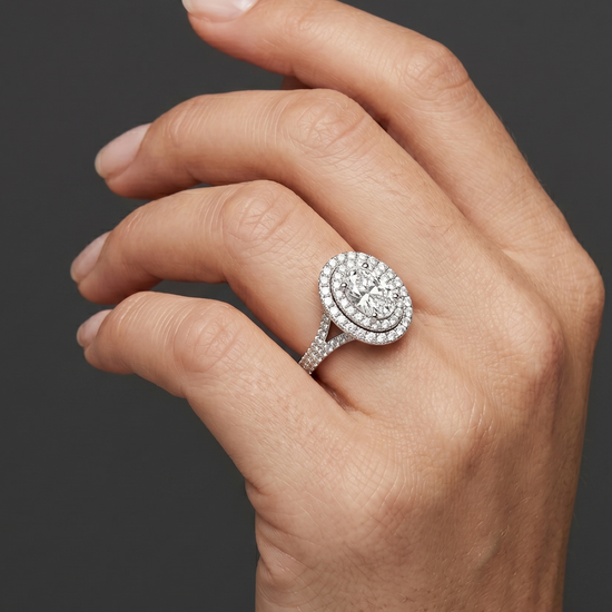 A close-up of a woman's elegantly posed hand wearing a brilliant oval-cut diamond ring with a double halo and a diamond-encrusted split-shank band, set against a smooth, dark charcoal gray background.