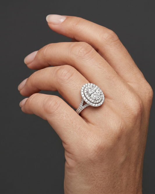 A close-up of a woman's elegantly posed hand wearing a brilliant oval-cut diamond ring with a double halo and a diamond-encrusted split-shank band, set against a smooth, dark charcoal gray background.
