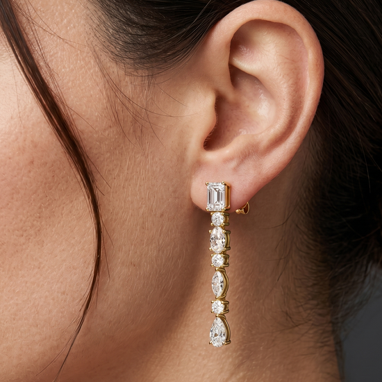 A close-up editorial photo of a woman's ear and jawline against a smooth, dark charcoal gray background. She is wearing a delicate, dangling yellow gold earring featuring a vertical row of clear, sparkling gemstones in alternating shapes. The design starts with an emerald-cut stud at the earlobe, followed by alternating round and marquise-cut stones, and ends with a pear-shaped drop. The woman has fair skin with natural texture and dark brown hair pulled softly back.