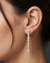 A close-up editorial photo of a woman's ear and jawline against a smooth, dark charcoal gray background. She is wearing a delicate, dangling yellow gold earring featuring a vertical row of clear, sparkling gemstones in alternating shapes. The design starts with an emerald-cut stud at the earlobe, followed by alternating round and marquise-cut stones, and ends with a pear-shaped drop. The woman has fair skin with natural texture and dark brown hair pulled softly back.