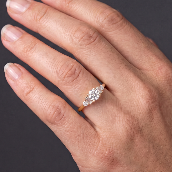 A close-up of a person's hand displaying a delicate gold ring. The ring has a round center diamond with smaller baguette and round side stones, set against a dark gray background.