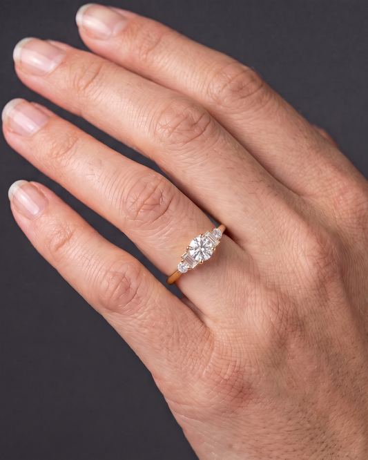 A close-up of a person's hand displaying a delicate gold ring. The ring has a round center diamond with smaller baguette and round side stones, set against a dark gray background.