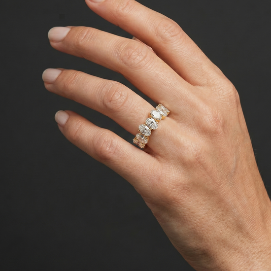 Close-up editorial photograph of an elegant woman's hand wearing a yellow gold ring set with a continuous row of oval-cut diamonds. The hand is posed gracefully against a smooth, dark charcoal gray background, with soft studio lighting illuminating the natural skin texture and the brilliance of the gemstones.