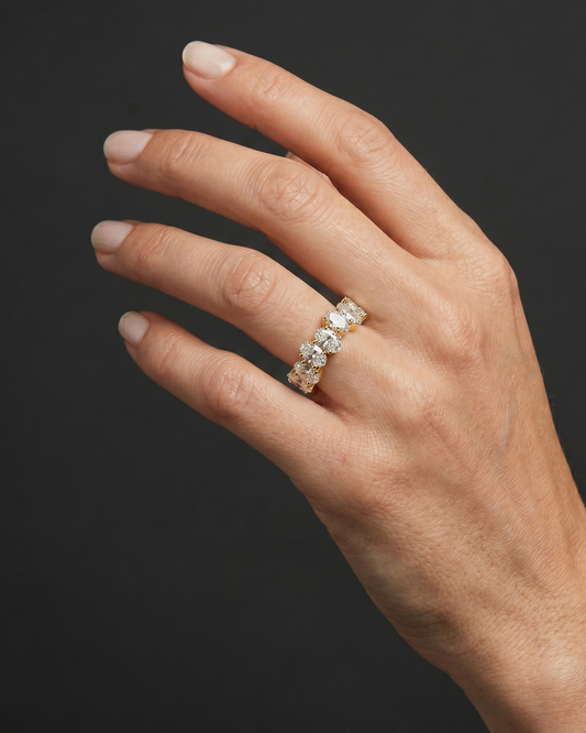 Close-up editorial photograph of an elegant woman's hand wearing a yellow gold ring set with a continuous row of oval-cut diamonds. The hand is posed gracefully against a smooth, dark charcoal gray background, with soft studio lighting illuminating the natural skin texture and the brilliance of the gemstones.