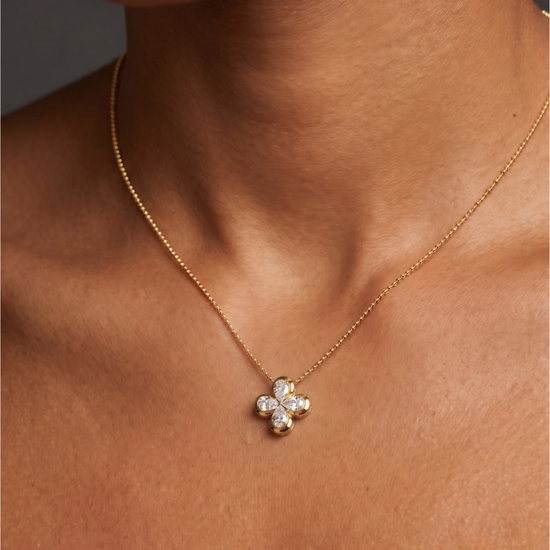Close-up of a person's neck and collarbone wearing a delicate gold chain necklace with a small, gold, four-petal floral or clover pendant featuring diamond accents.