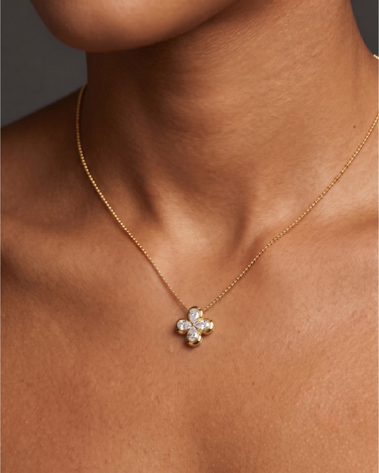 Close-up of a person's neck and collarbone wearing a delicate gold chain necklace with a small, gold, four-petal floral or clover pendant featuring diamond accents.