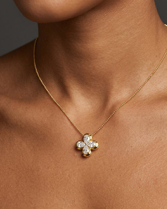 Close-up of a person's neck and collarbone wearing a delicate gold chain necklace with a small, gold, four-petal floral or clover pendant featuring diamond accents.