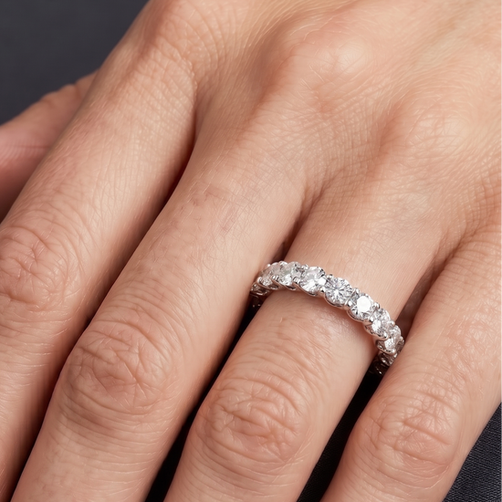 Close-up of a woman's hand resting gracefully against a dark charcoal background, showcasing a luxury diamond eternity band on her ring finger. The silver-toned ring features a continuous row of round-cut diamonds held in a distinctive twisted, woven prong setting. 