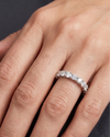 Close-up of a woman's hand resting gracefully against a dark charcoal background, showcasing a luxury diamond eternity band on her ring finger. The silver-toned ring features a continuous row of round-cut diamonds held in a distinctive twisted, woven prong setting. 