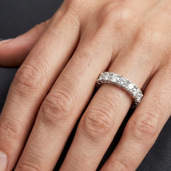 Close-up of a woman's hand resting gracefully against a dark charcoal background, showcasing a luxury diamond eternity band on her ring finger. The silver-toned ring features a continuous row of round-cut diamonds held in a distinctive twisted, woven prong setting. Soft, diffused lighting highlights the brilliance of the gems and the natural texture of her skin.