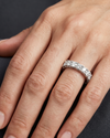 Close-up of a woman's hand resting gracefully against a dark charcoal background, showcasing a luxury diamond eternity band on her ring finger. The silver-toned ring features a continuous row of round-cut diamonds held in a distinctive twisted, woven prong setting. Soft, diffused lighting highlights the brilliance of the gems and the natural texture of her skin.